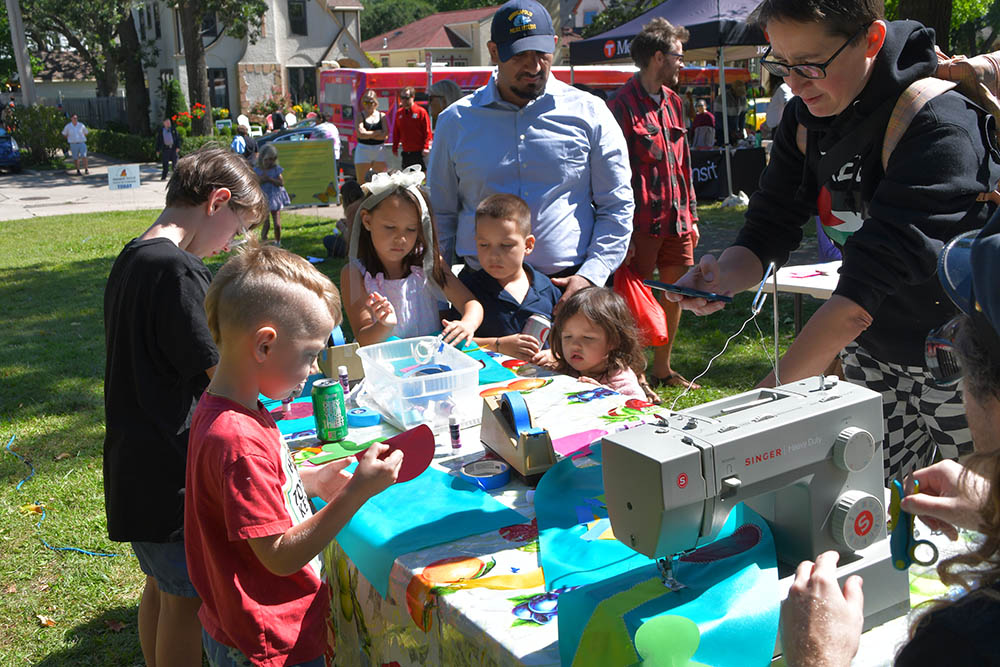 Children choose from a variety of shapes and colors of fabrics to create flags representing the most important things about regional parks. The flags are being sewn by an assistant to artist Peter Haakon Thompson (not pictured). At table, children choose shapes to place on bright blue flags and place them with painter’s tape to the fabric. A man sews the flags on a sewing machine at the end of the table.