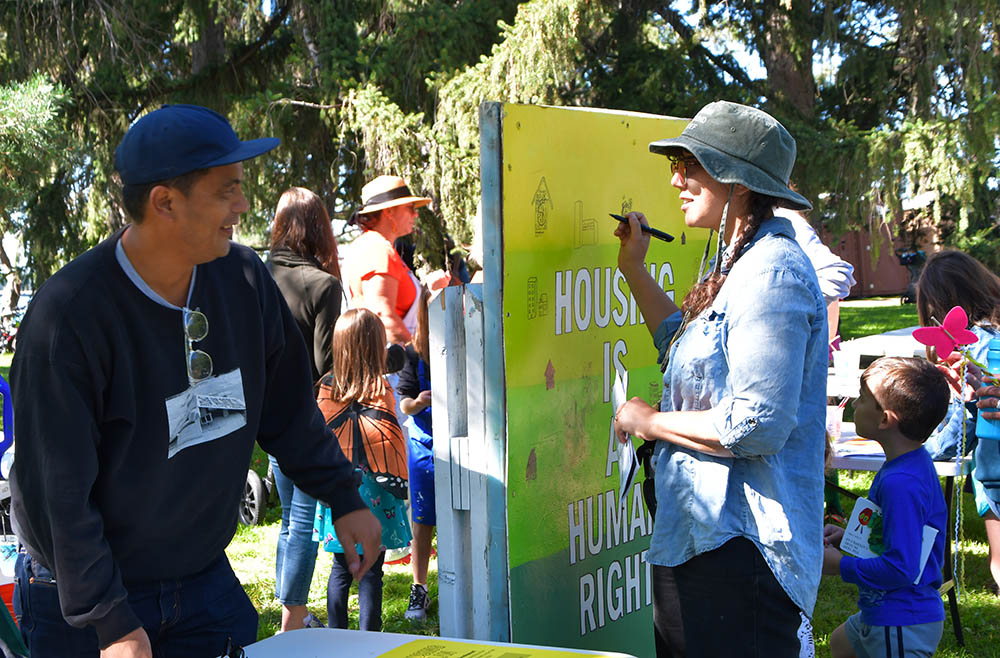 Artist Will Siasoco (left) designed a large poster (right) on which he encouraged people to draw what housing looks like to them. Man in baseball cap talks with woman using felt-tip pen to draw on multi-color poster that says, “Housing is a human right.”