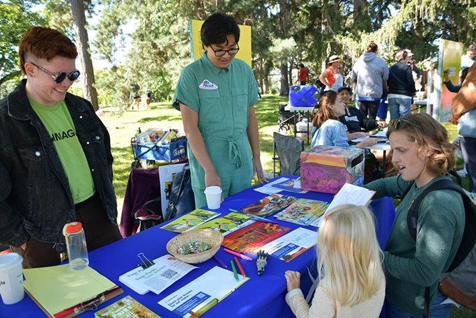 Visitors to the annual Monarch Festival at Nokomis-Hiawatha Regional Park shared their dreams for the future of the region on colorful postcards with designs by four visual artists for the Art+Policy project. Woman and child at table staffed by two Met Council employees fill out a postcard.