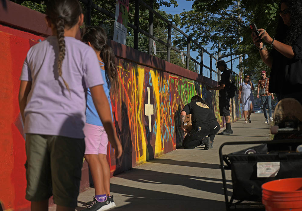 Visitors to the Chromazone Mural & Art Festival in Saint Paul paint a mural designed by community members and Thomasina Topbear, Oglala Lakota and Santee Sioux artist (not pictured). Two men and one child paint on a long mural, about four feet high, next to a sidewalk in the Raymond/University Avenue area of Saint Paul.