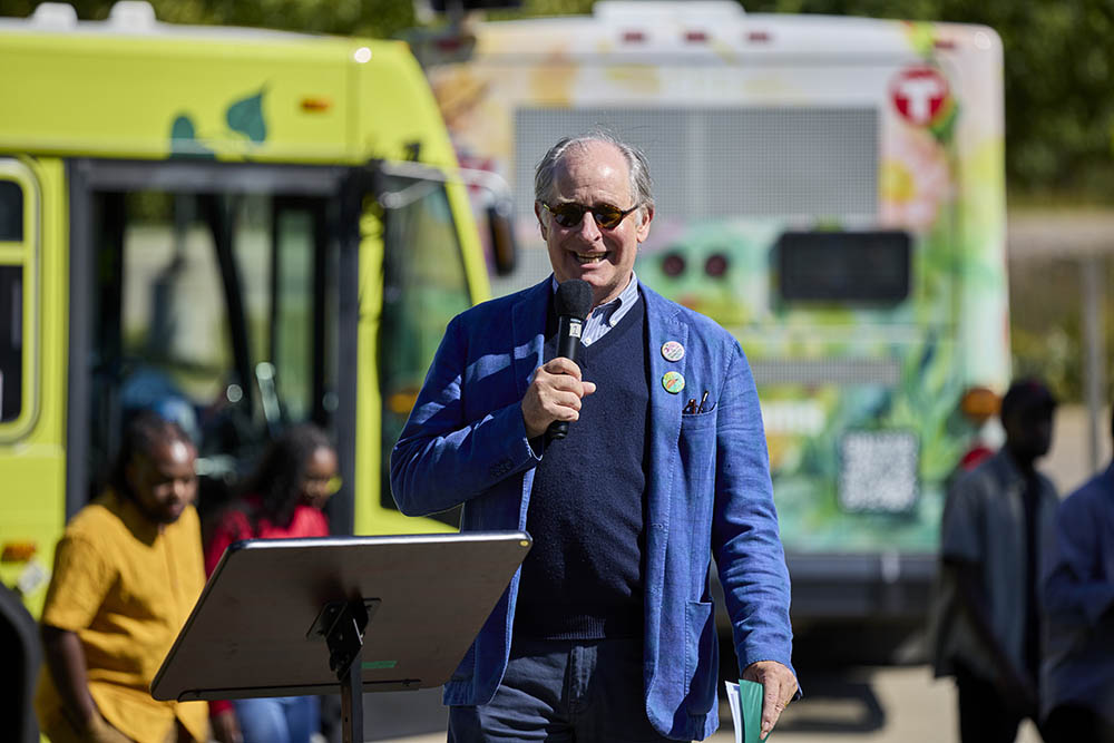 Charlie Zelle, Chair of the Met Council, speaks at the “Art, Policy, Pizza & Ping Pong” event at Lebanon Hills Regional Park to a backdrop of Metro Transit buses wrapped in colorful designs. Charlie Zelle stands with microphone in front of two colorful Metro Transit buses.