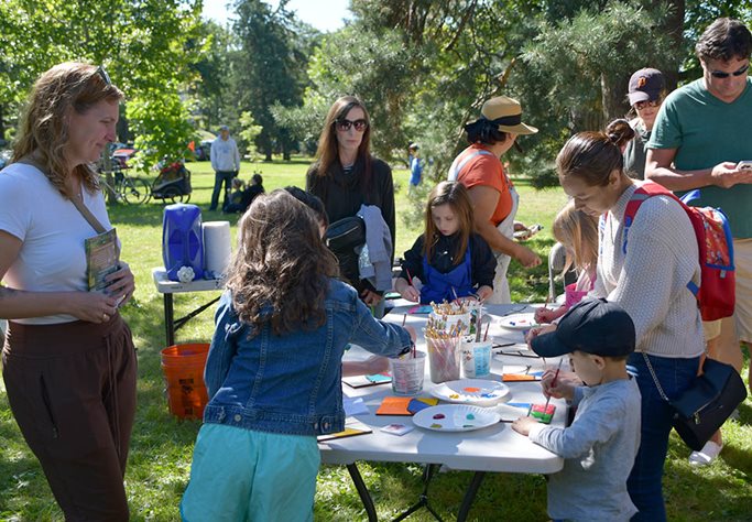 Children and adults paint predesigned tiles for assembly into three large visual art pieces depicting people’s dreams for the future of the region. People stand around a table with paints and paintbrushes, painting tiles in bright colors.
