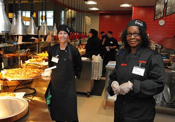 Two women pause from baking and serving pizza.