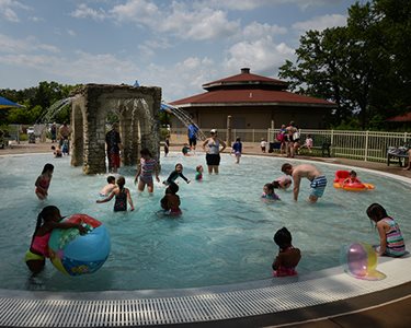 Children wade and splash in shallow pool with central water fountain.