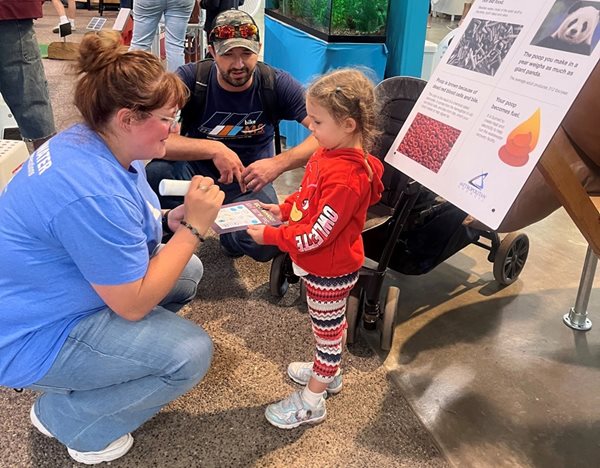 A volunteer squats down to mark the bingo card carried by a child, with an adult in the background, that shows they visited the exhibit.